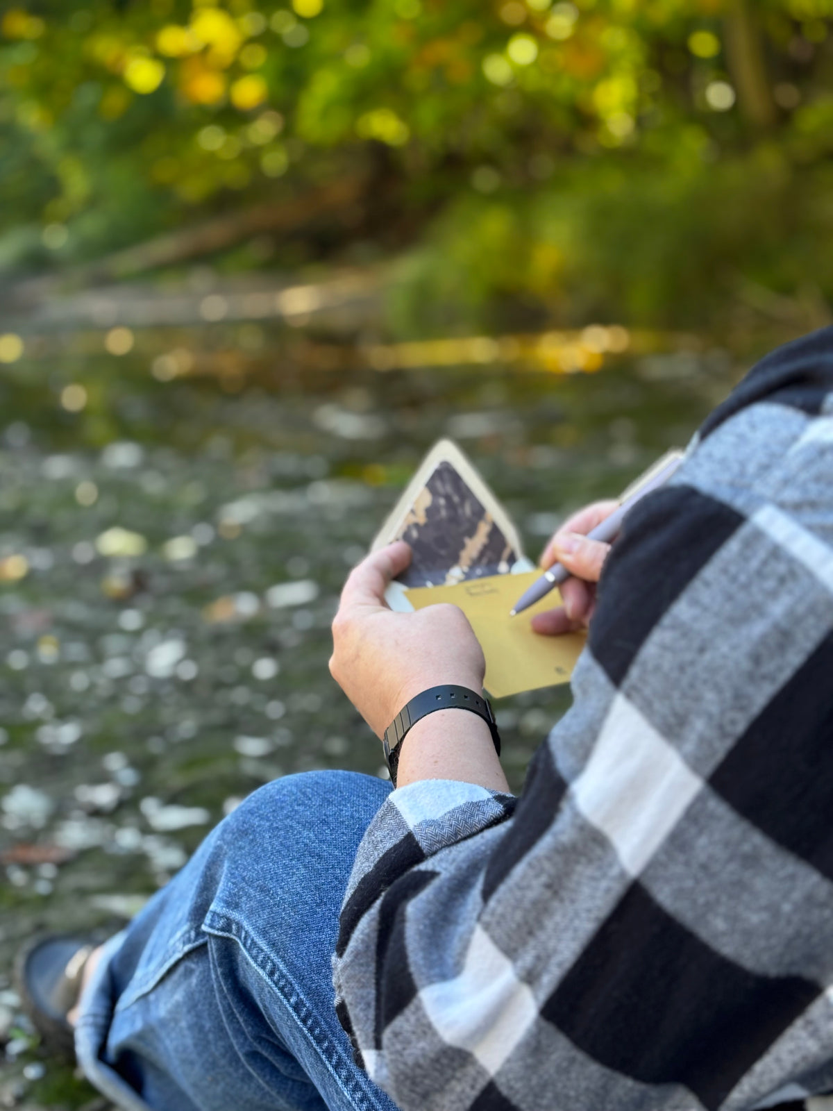 Person writing on a golden yellow Gneiss notecard beside water, with its rock-lined envelope in hand.