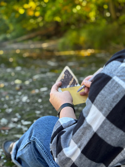 Person writing on a golden yellow Gneiss notecard beside water, with its rock-lined envelope in hand.