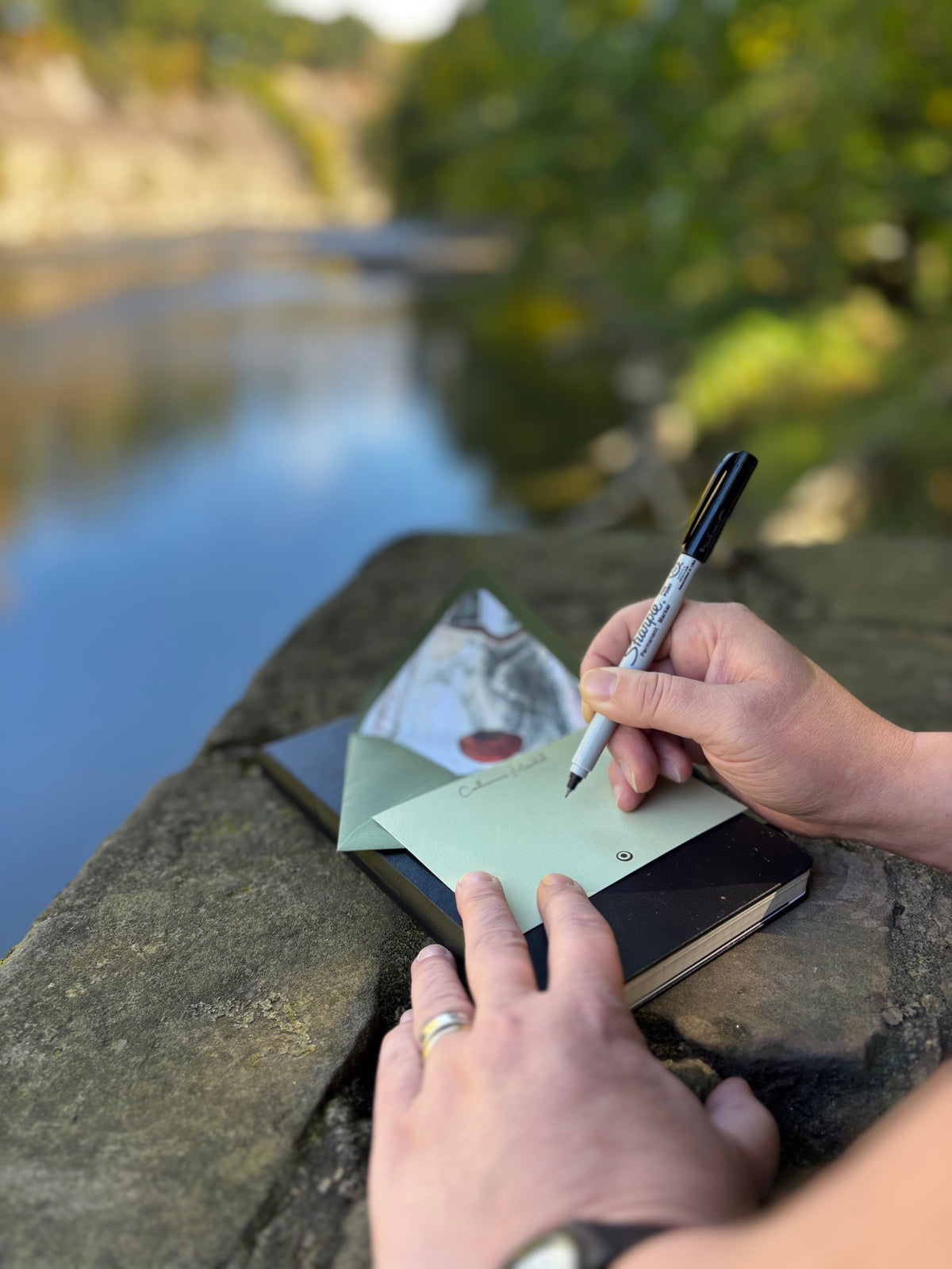 Person writing on a pale green Half Shell notecard beside water, with its oyster-lined envelope behind it.