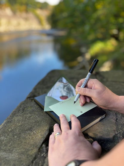 Person writing on a pale green Half Shell notecard beside water, with its oyster-lined envelope behind it.