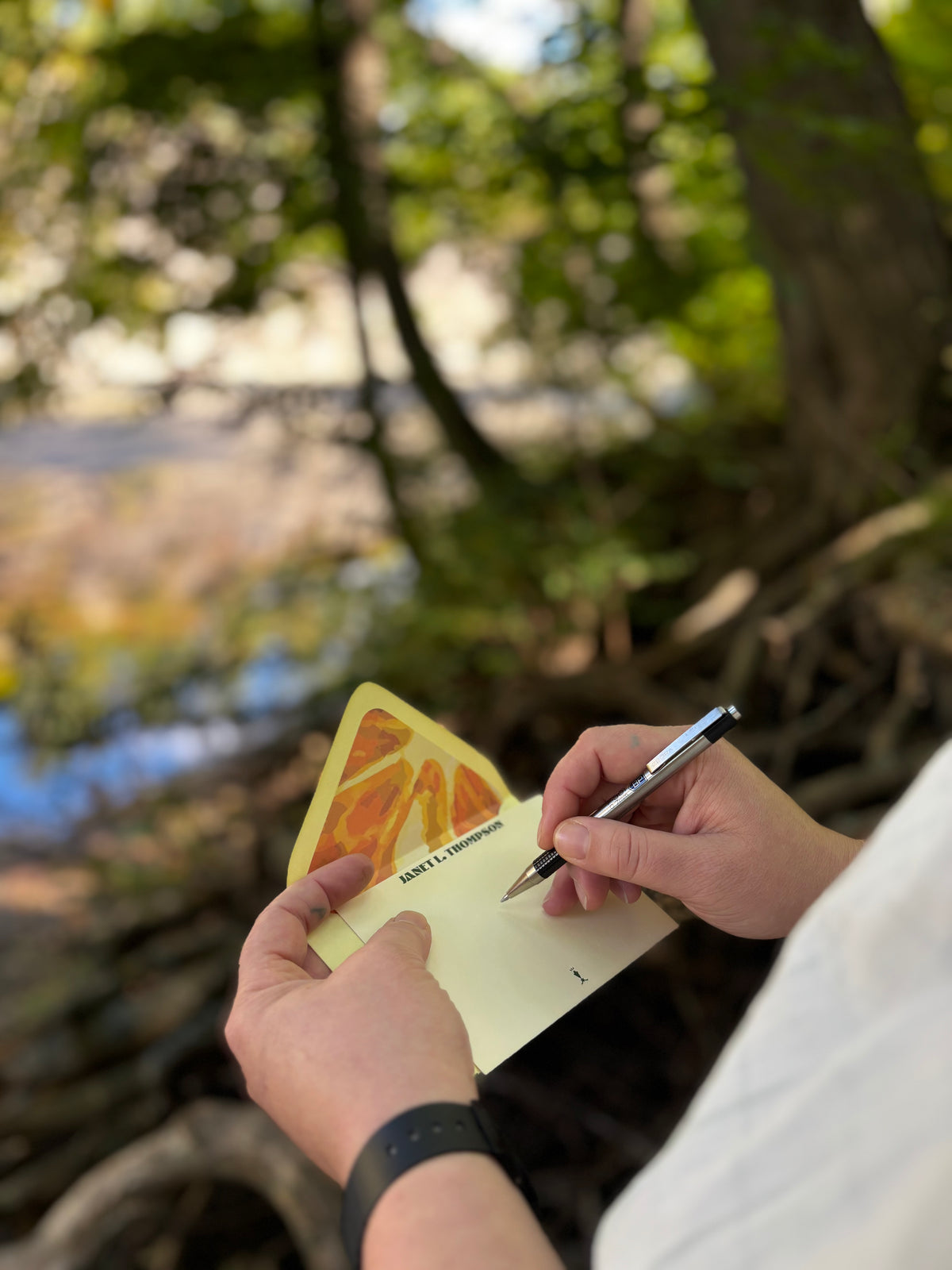 Person writing on an Orange You Glad cream notecard beside water, with an orange slice-lined envelope behind it.