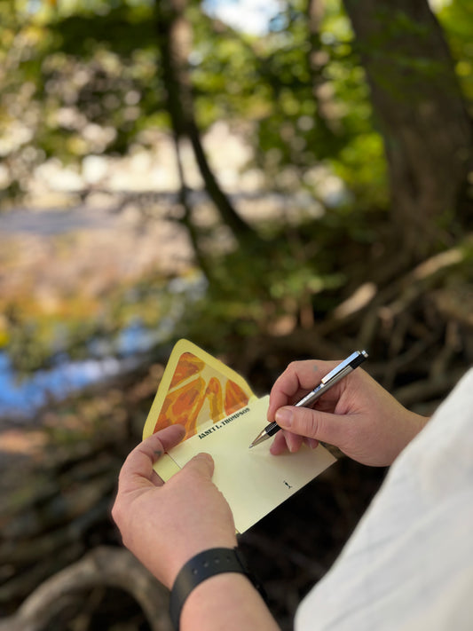 Person writing on an Orange You Glad cream notecard beside water, with an orange slice-lined envelope behind it.