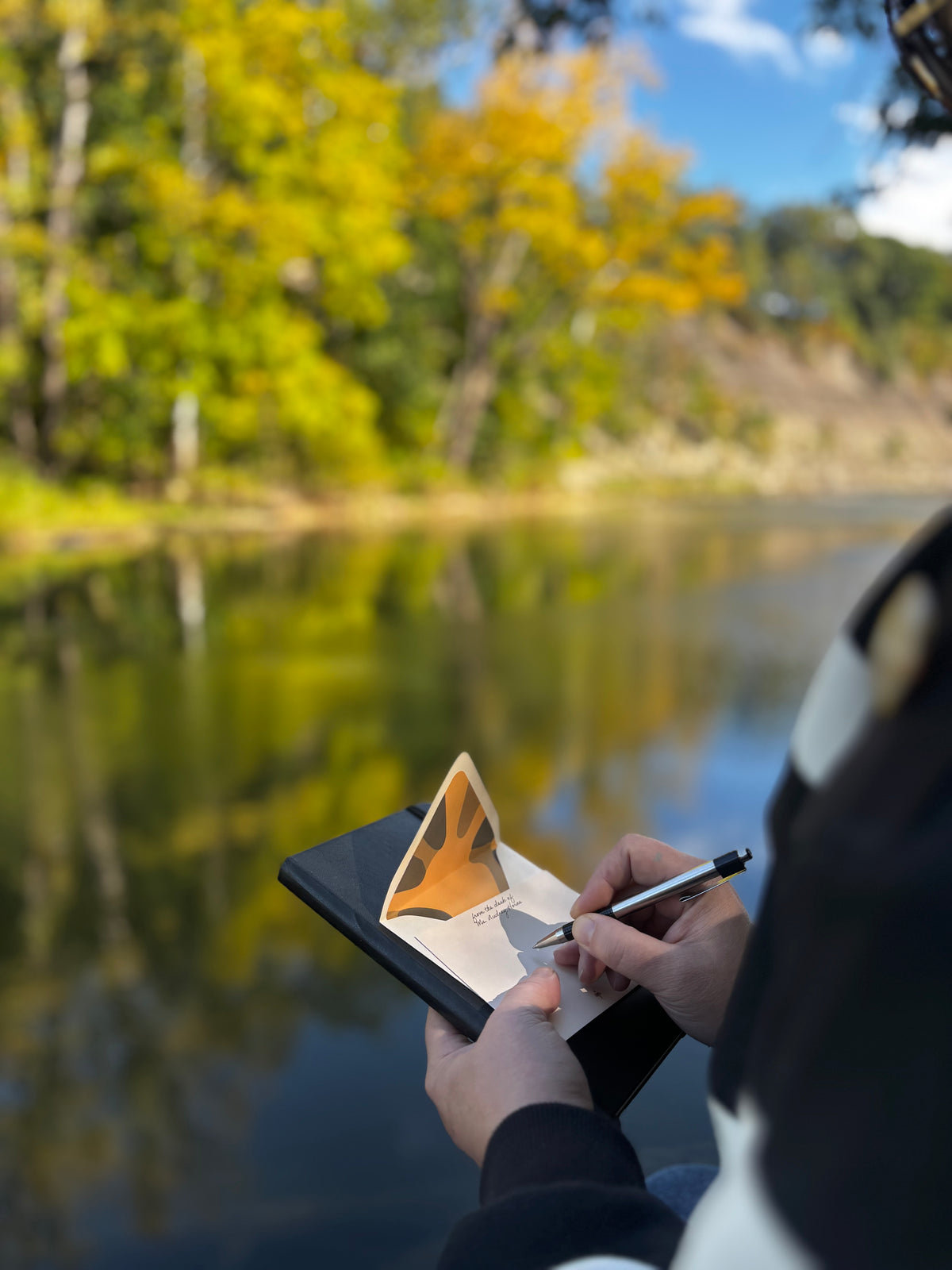 Person writing on a cream Poppy notecard beside water, with its lined envelope behind it.