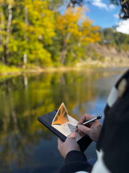 Person writing on a cream Poppy notecard beside water, with its lined envelope behind it.