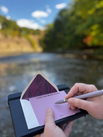 Person writing on a dilute wine-colored Meandering River notecard beside a river, with its lined envelope behind it.
