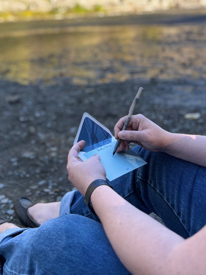 Person writing on a pale blue Fall Line notecard outdoors beside water, with its blue-lined envelope behind it.